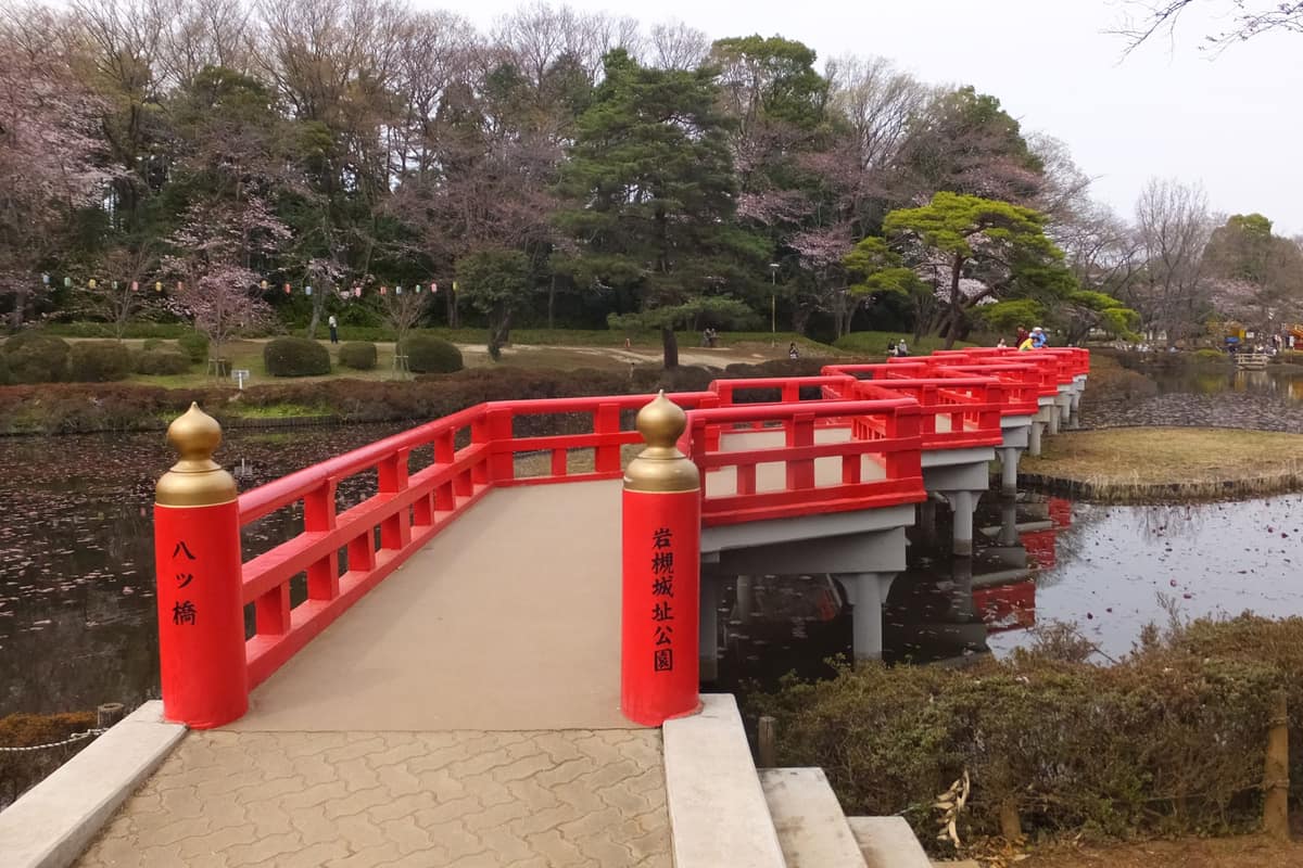 The vermillion Yatsuhashi (Eight Bridges) walkway in Iwatsuki Castle Ruins Park, with the park's name carved on the bridge post