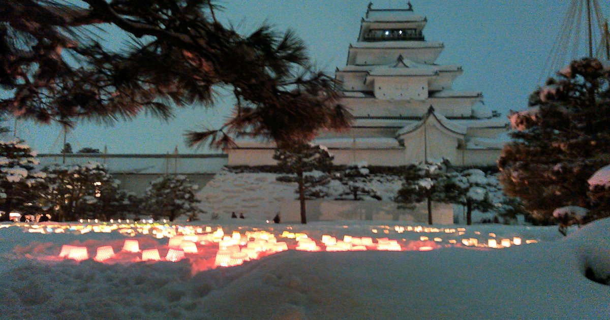 繪蠟燭祭晚上的鶴城：積雪的空地上排著一大片點亮的燭火，後方是屋頂積雪的天守閣，左上垂著松枝