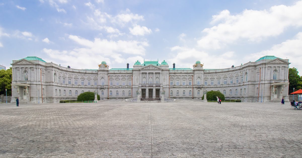 從前庭正中央看迎賓館赤坂離宮本館正面:灰白色石造宮殿左右對稱,兩翼向前彎成弧形,屋頂是綠色銅板,中央三角楣下有正面玄關,前面是一大片鋪石廣場,藍天白雲,遠處有零星幾位遊客