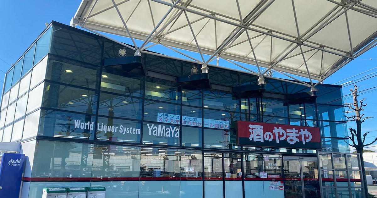 A Japanese discount liquor store, Sake no Yamaya: a glass-fronted box building under a white tensile-fabric canopy against a blue sky, white lettering reading World Liquor System YaMaYa across the glazing, a large red sign reading 酒のやまや to the right, and opening hours of 10:00 to 21:00 posted on the entrance door.