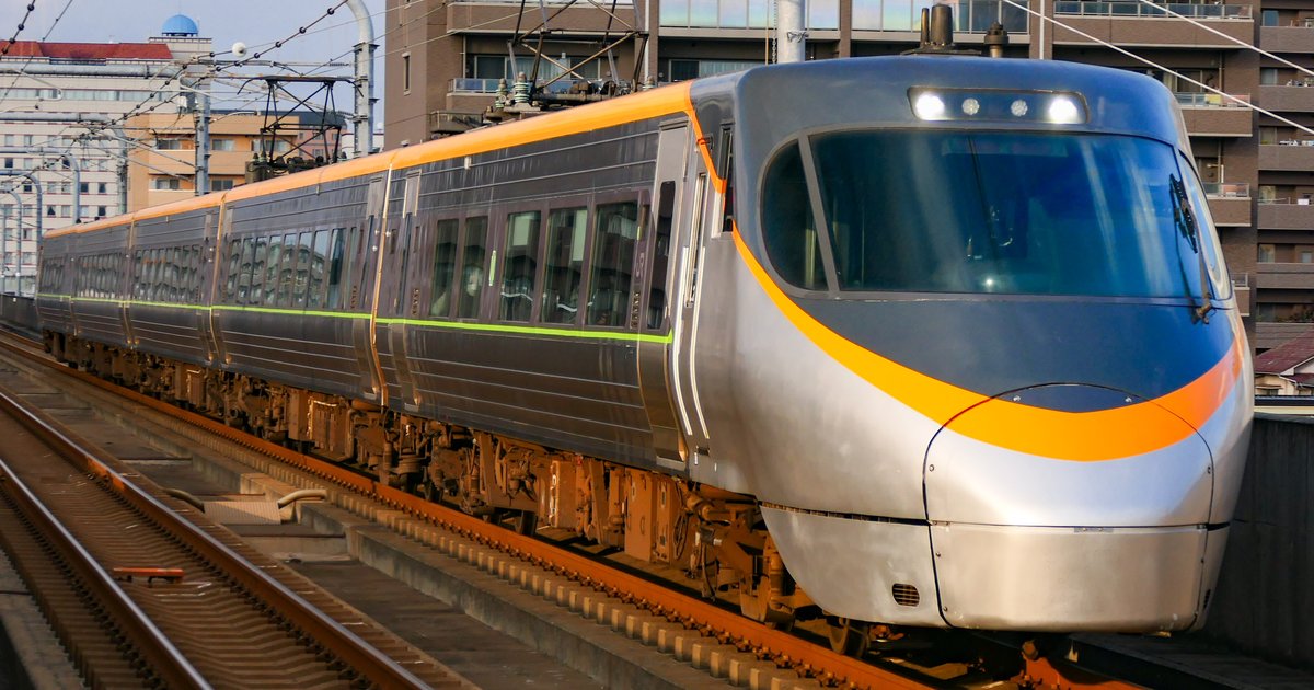 JR 四國 8000 系特急潮風號朝右前方駛來,銀色流線型車頭配橘色飾帶,車身側面是深灰色車窗與一條黃綠色細線,背景是高架旁的公寓大樓與電車架線