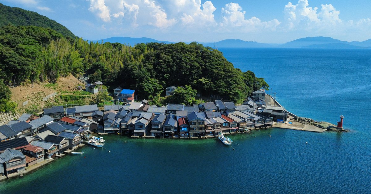 Aerial view of the Ine funaya, a row of timber houses whose ground floors sit right at the waterline with fishing boats moored in front and a red lighthouse at the end of the breakwater