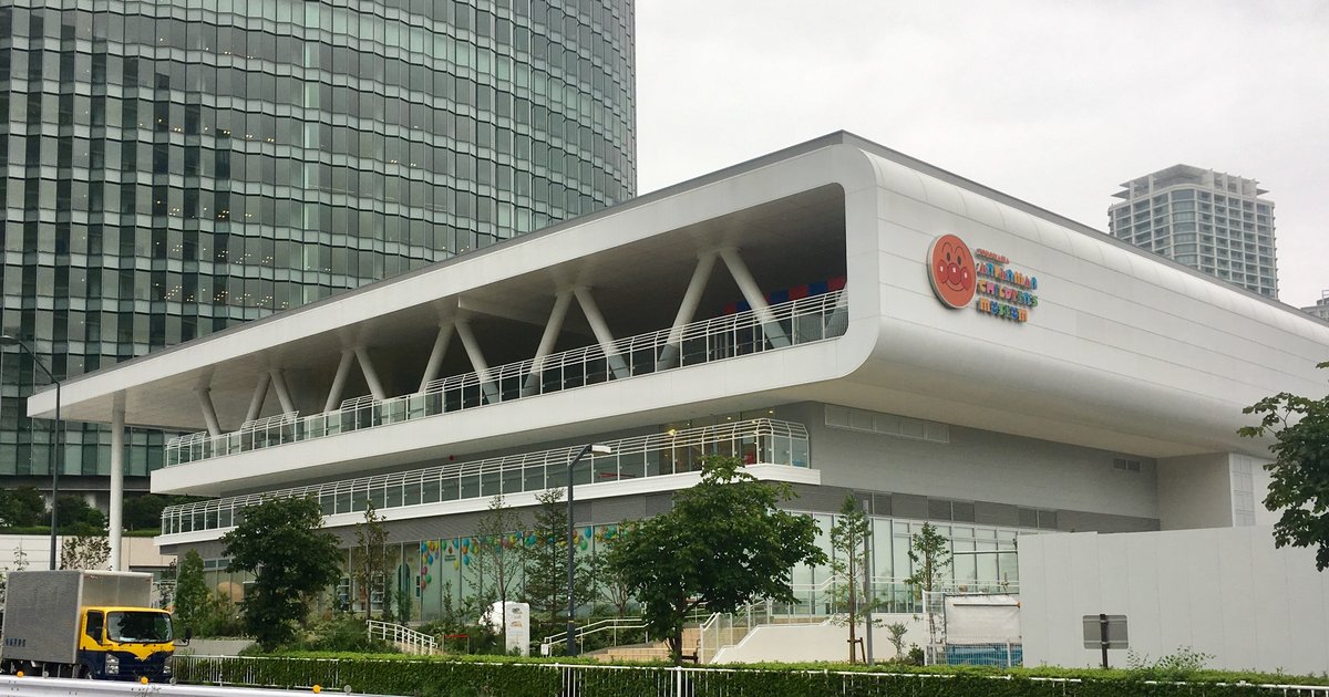 陰天從馬路對面看橫濱麵包超人兒童博物館，白色長方形建築有層層外推的露台與斜撐柱，牆上掛著圓形麵包超人臉標誌與彩色館名，左後方是玻璃帷幕高樓，前景是護欄與綠籬