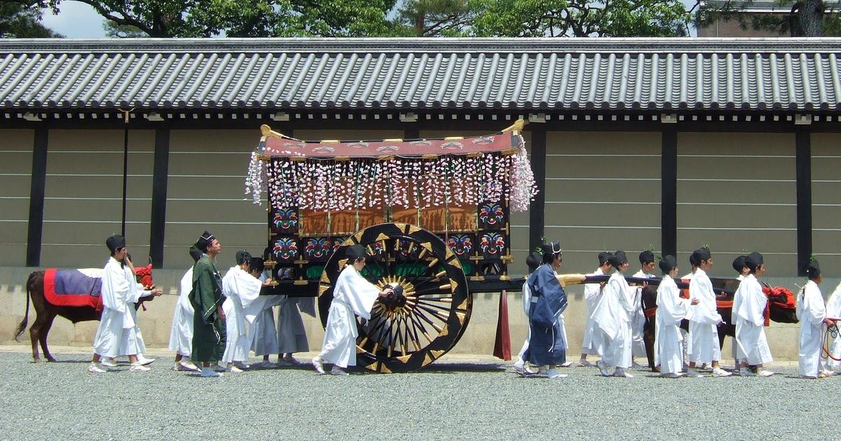 葵祭的牛車(御所車)行經京都御所的築地塀前:黑漆大車輪、車篷前垂著整排藤花裝飾,前方一頭披紅色鞍布的牛,周圍是穿白色與藍、綠色平安裝束的隨從,地面是京都御苑的碎石