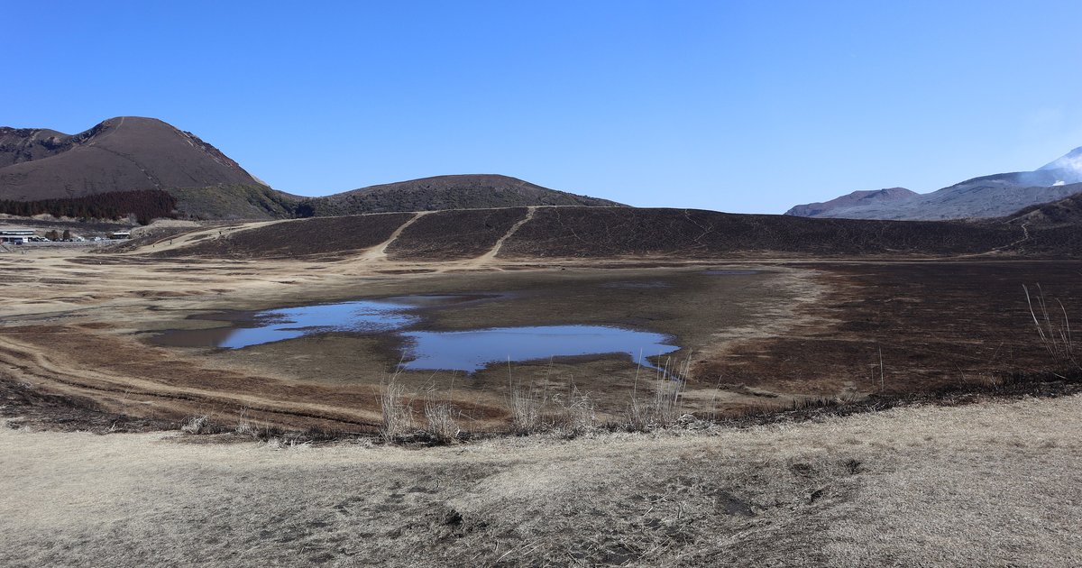 野燒後的阿蘇草千里ヶ浜：中央兩個淺水窪，後方山坡被燒成一整片深褐色、留著幾道淺色步道，左後方是另一座深褐色的火山丘，右邊遠處是冒白煙的中岳