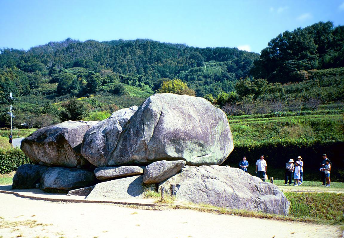 Ishibutai Kofun: two enormous grey granite boulders resting on lower stones with the mouth of a burial chamber between them, a sandy path in front, a small group of visitors standing at the right for scale, and terraced green fields below a forested hillside behind