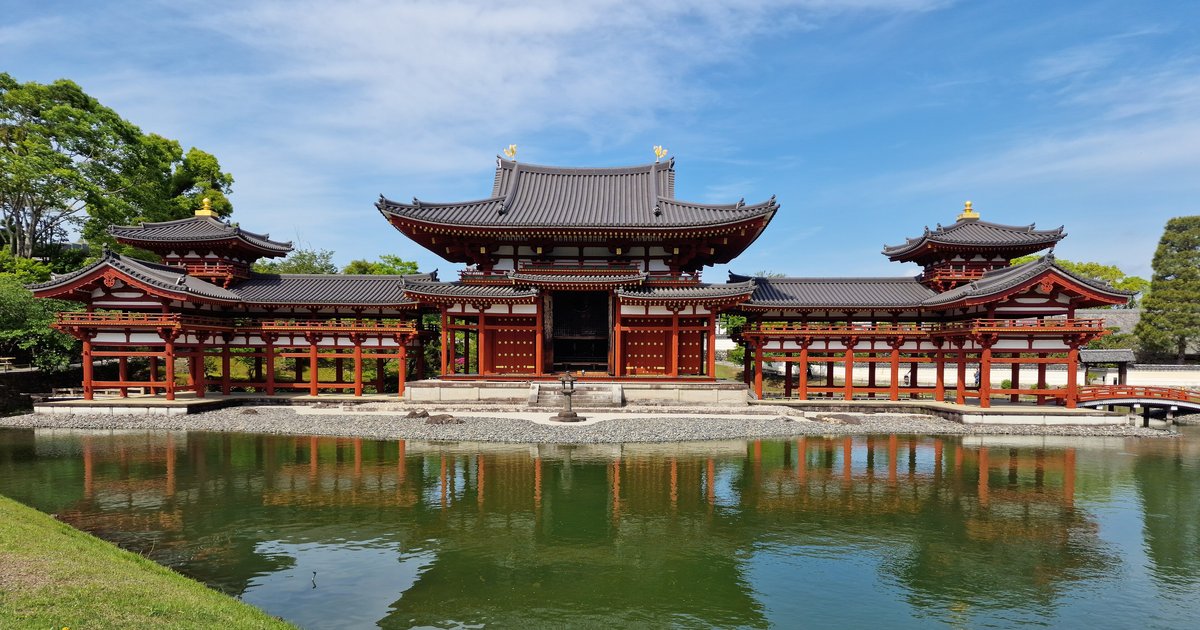 晴天從阿字池對岸正面看宇治平等院鳳凰堂:中堂與左右兩座翼廊對稱展開,朱紅柱子倒映在綠色池面,中堂屋脊上一對金色鳳凰,畫面裡沒有遊客
