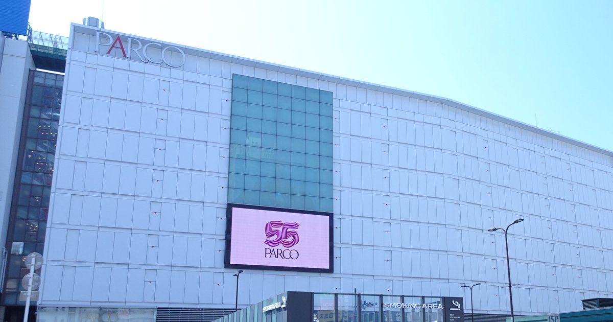 池袋 PARCO 本館的白色外牆,左上角掛著 PARCO 字樣,中央是一片綠色玻璃帷幕,下方粉紅色大螢幕顯示 PARCO 標誌,晴天