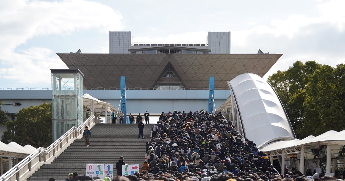 冬季 Comiket 入場日的東京 Big Sight:逆三角形的會議棟矗立在正中央,下方寬大的石階上擠滿穿深色厚外套的入場人潮,從畫面下緣一路往上走向會場,右邊是白色帆布造型的天橋屋頂與綠樹