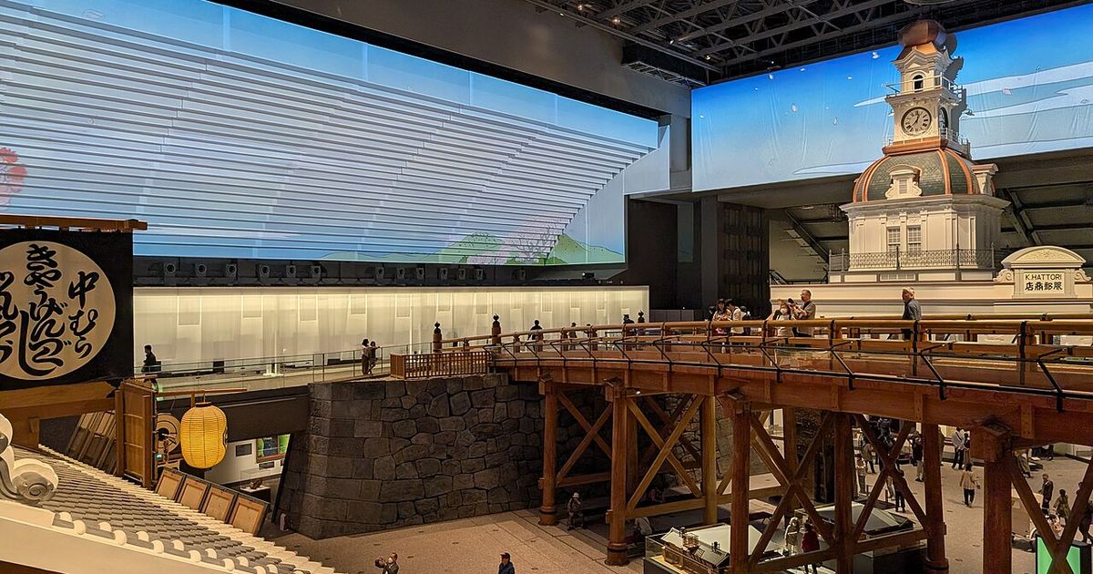 Sixth-floor permanent exhibition of the Edo-Tokyo Museum: visitors walking across the timber reconstruction of Nihonbashi, with the white Hattori clock tower model on the right and a wall-sized blue sky screen behind