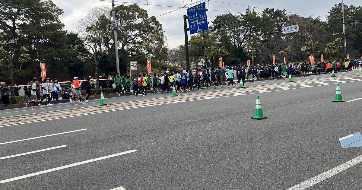 陰天下的愛媛馬拉松:一群穿彩色衣服的跑者沿著電車軌道旁的大路跑過,路邊立著「42 km」距離牌與橘色旗,綠色背心的志工與觀眾站在松樹下的人行道旁,路面擺著綠色三角錐
