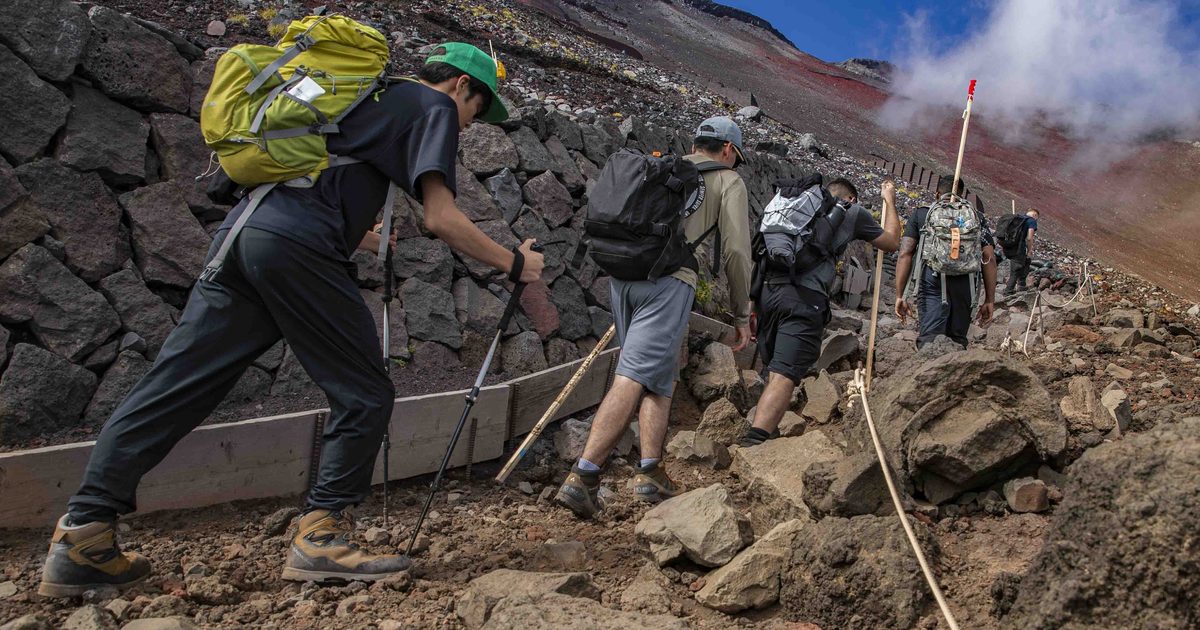 A line of hikers with large packs, trekking poles and wooden staffs climbs a steep rocky pitch beside a dry-stone retaining wall on Mount Fuji, a thick guide rope strung along the right