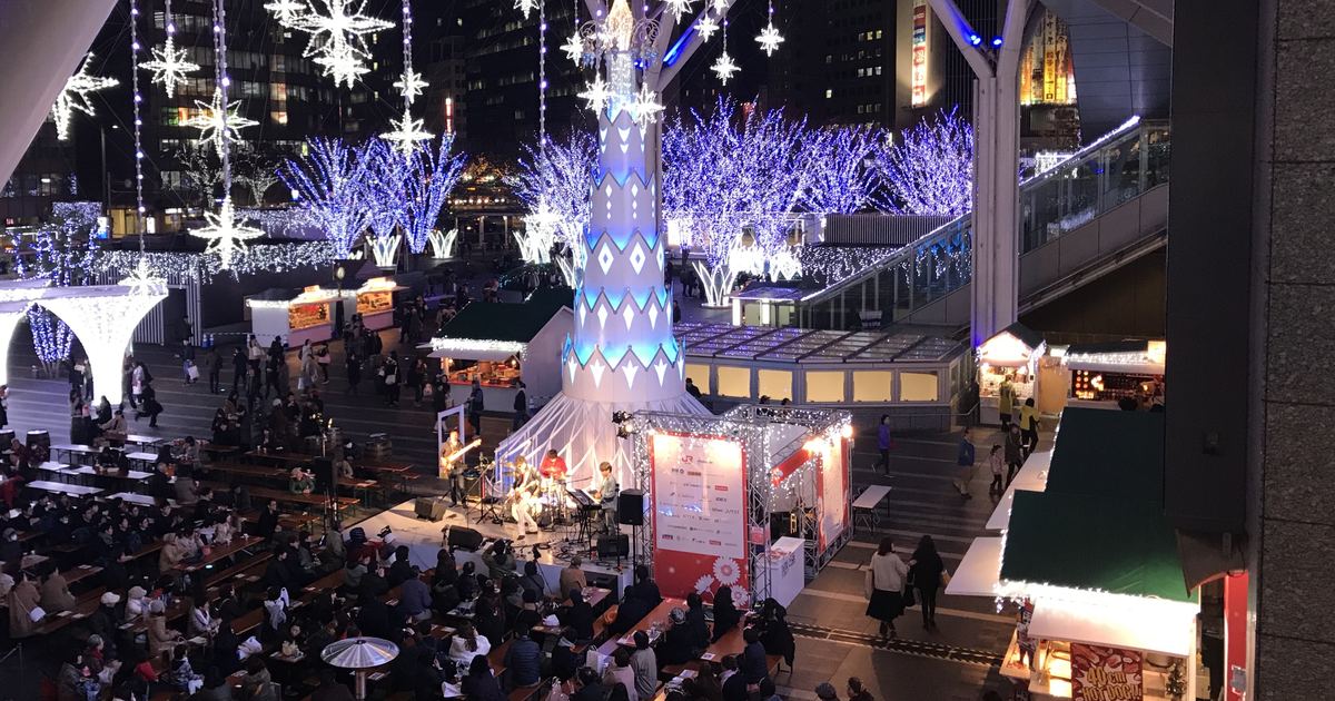 2018 年 12 月從高處俯看 JR 博多站前廣場的聖誕市集夜景：中央一座白藍相間的塔形聖誕樹，樹下是有樂團演出的舞台，左下坐滿人的長桌區，四周是亮著暖黃燈的木屋攤位，後方一整排纏著藍白燈的行道樹，頭頂垂掛星形燈飾