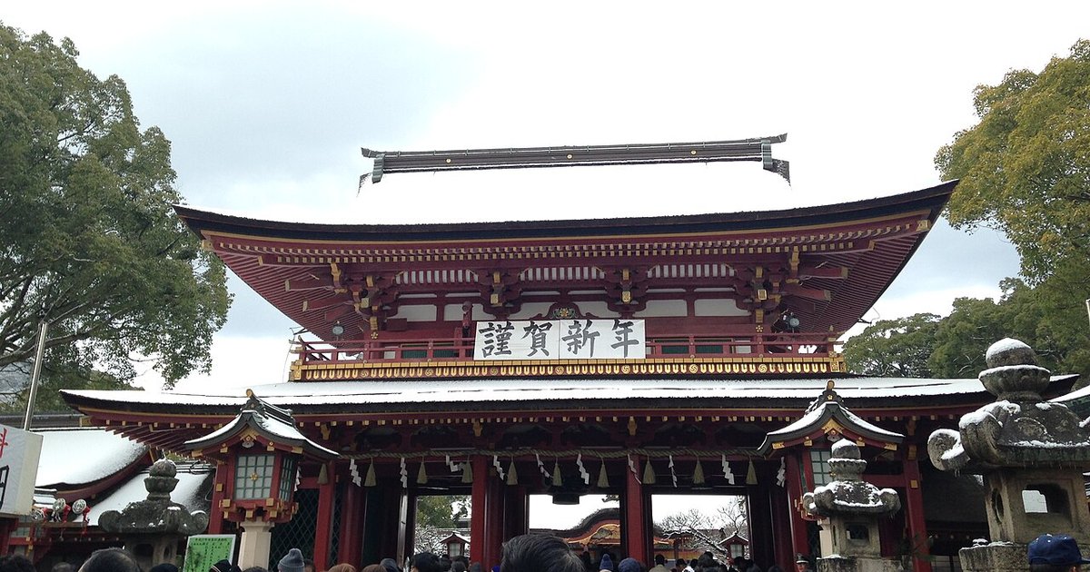 2015 年元旦的太宰府天滿宮樓門：朱紅色的兩層門樓，二樓欄杆上掛著白底黑字「謹賀新年」橫幅，屋頂與兩側石燈籠上有積雪，門前擠著初詣的人群