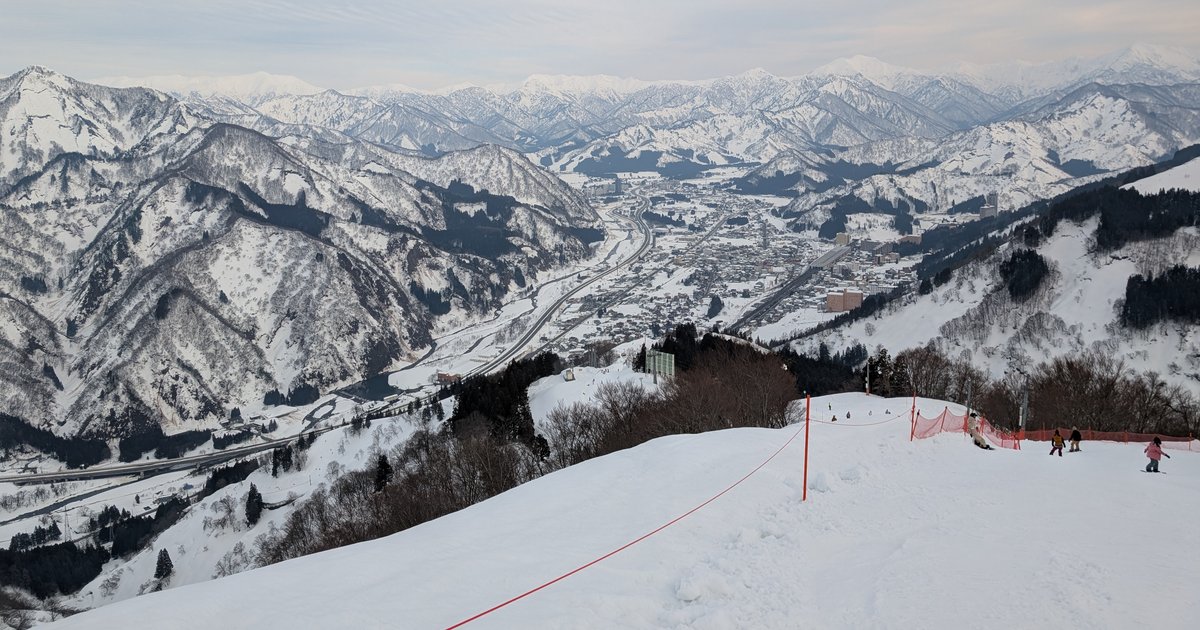 Looking down a groomed upper run at GALA Yuzawa: a red boundary rope and orange safety netting, with the town and expressway in the Yuzawa valley and snow-covered mountains beyond