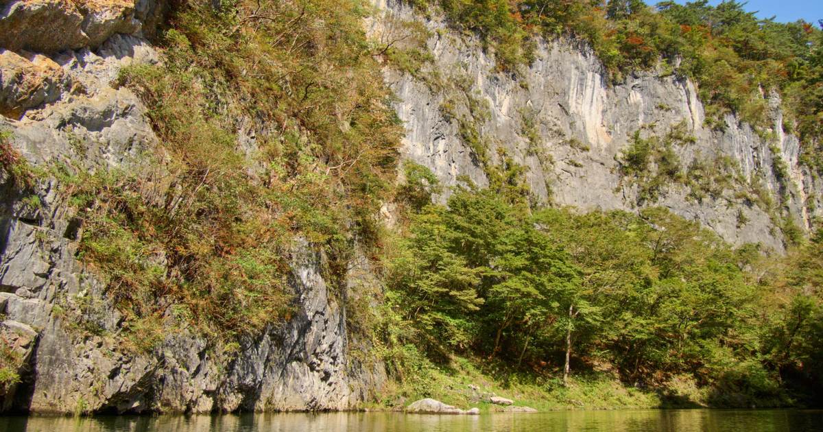 猊鼻溪的石灰岩壁:灰白色的岩壁從平靜的砂鐵川水面直直立起,岩壁上方與右側長著雜木林,葉子略帶黃褐色,頂上是藍天