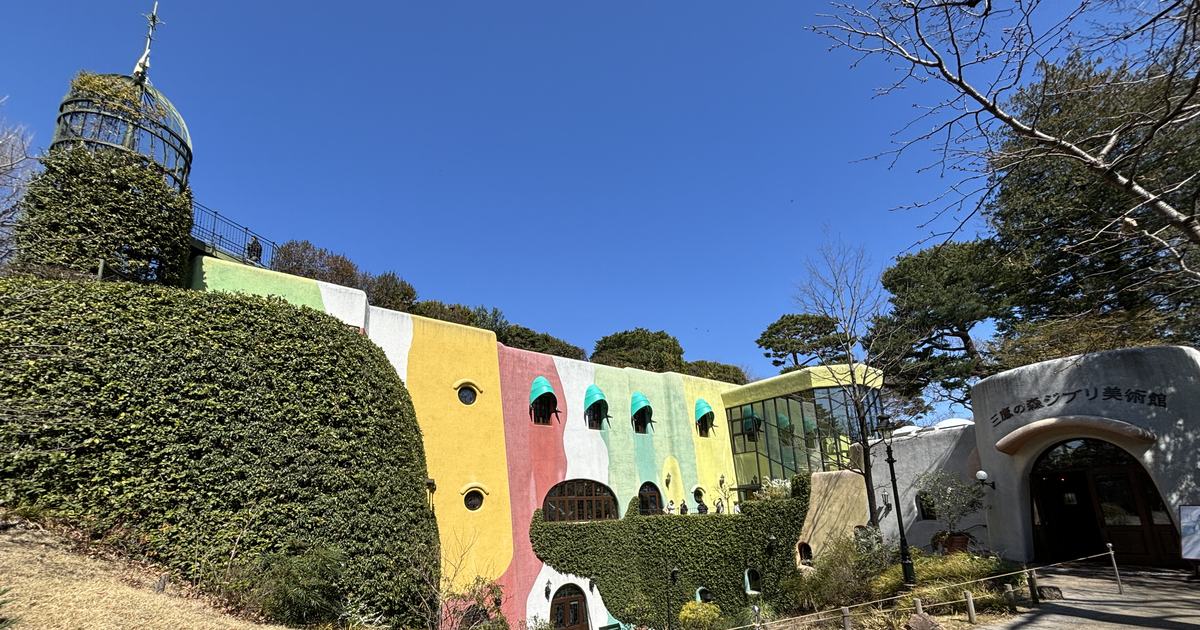 The museum exterior under a cloudless blue sky: the left half is a tall clipped ivy hedge topped by an ivy-covered round tower with a green metal domed cage; the right half is a stucco wall painted in irregular blocks of pale green, white, yellow, pink and cream, pierced by small round windows and four arched windows under turquoise awnings; at the far right a white curved archway forms the entrance, with a sign reading 三鷹の森ジブリ美術館 mounted above it, above a flagstone path and winter-brown grass