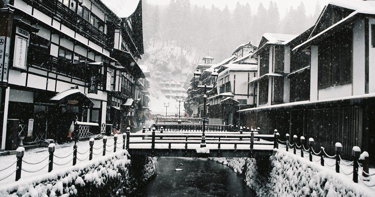 下雪中的銀山溫泉街:銀山川兩岸是三、四層樓的木造旅館,河上橫著一座木橋,岸邊的鐵鍊欄杆柱與煤氣燈都頂著一層雪,路面全是雪、看不到任何車輛