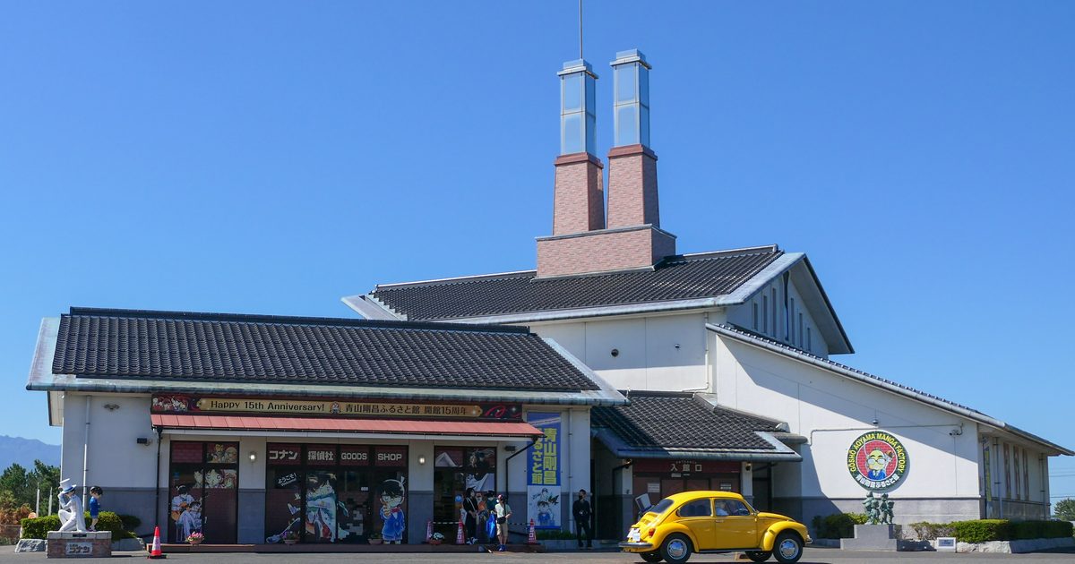 藍天下的青山剛昌故鄉館舊館,黑瓦白牆的建築頂上立著兩支磚紅色煙囪,左翼是掛著周年布條的周邊商店,右翼入館口前停著一輛黃色金龜車