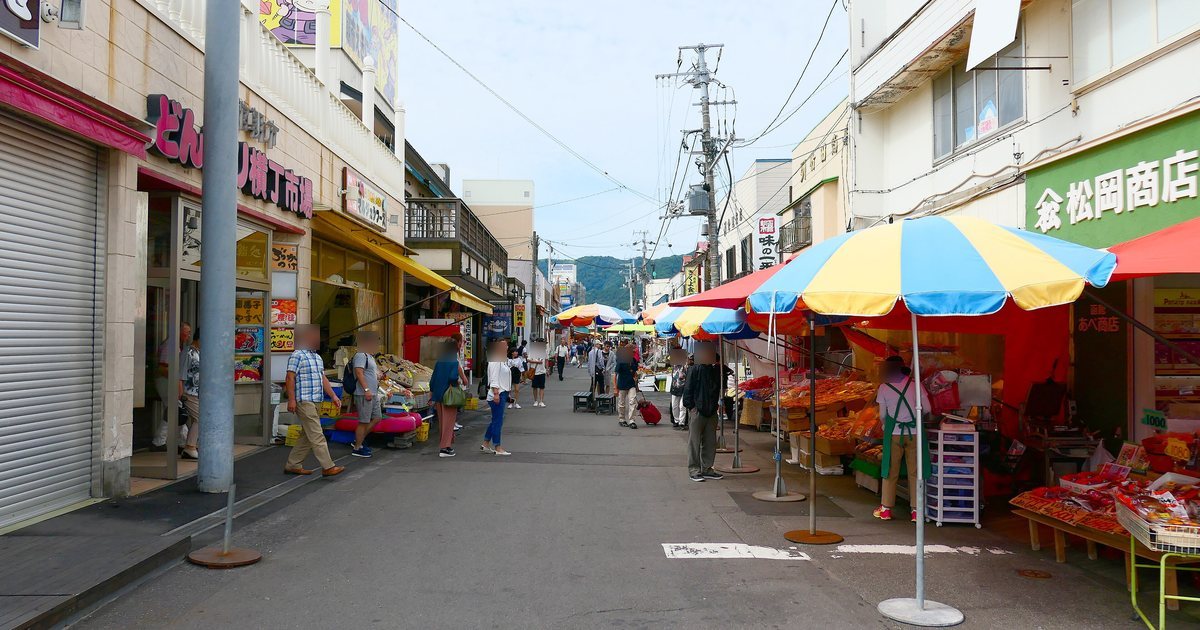 早上的函館朝市街道：左邊是掛著「どんぶり横丁市場」紅字招牌的建築，右邊一排藍黃相間的大陽傘下擺著海產攤，遊客沿著街往裡走，遠處是綠色的山