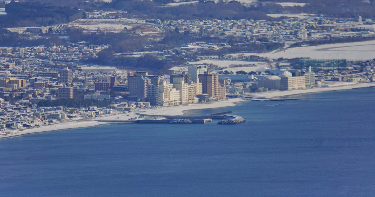 從函館山遠望冬天的湯之川溫泉一帶：深藍色的海面上方，一段積雪的白色沙灘沿著海岸彎過去，岸邊排著十幾棟米色與淺褐色的溫泉飯店大樓，前方有一道彎曲的防波堤，後方是覆雪的住宅區與山丘