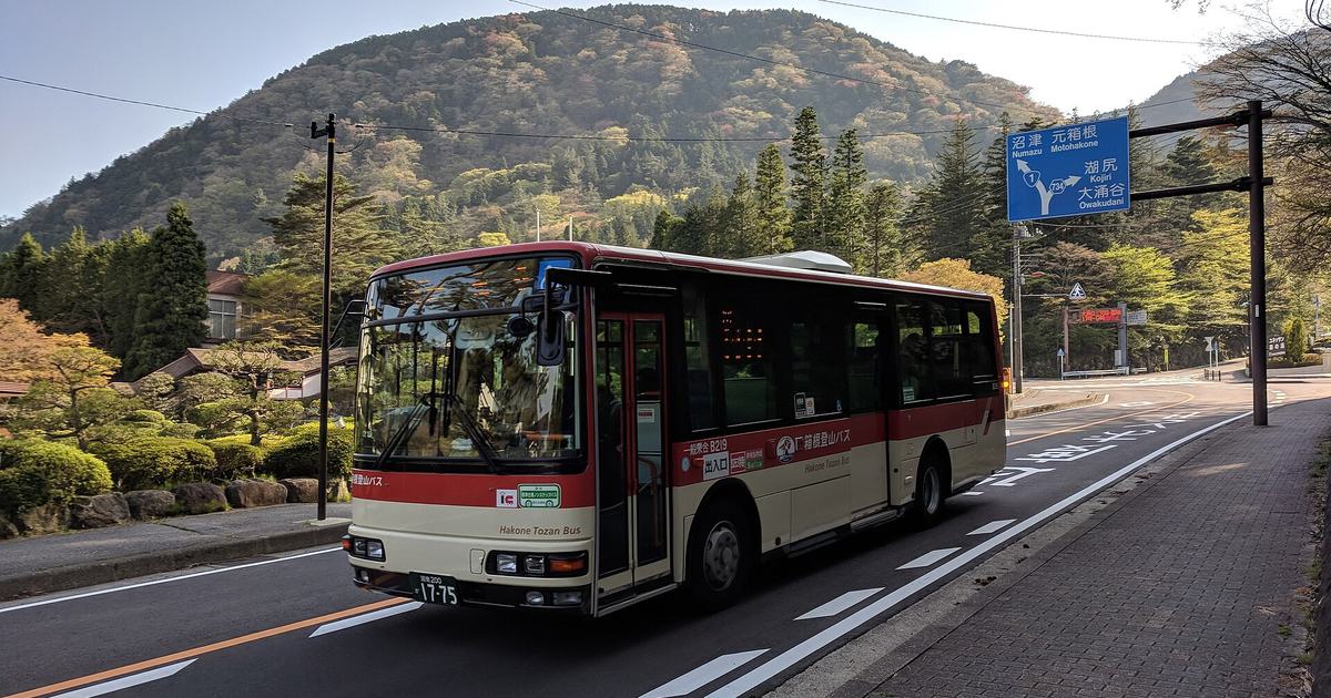 A red and cream Hakone Tozan Bus travelling along a two-lane road through the Hakone mountains, Hakone Tozan Bus lettered along its side, with a blue direction sign to the right showing Numazu and Motohakone beside a National Route 1 shield and Kojiri and Owakudani below, against a fully forested hillside