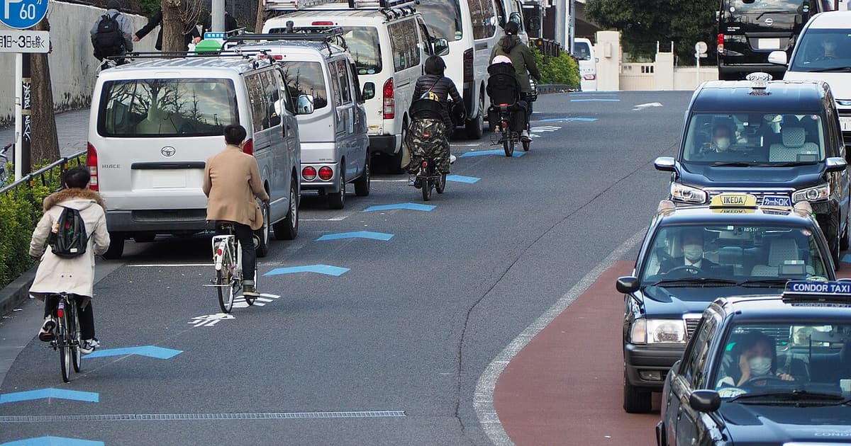 A street in Harajuku, Tokyo: four cyclists riding away from the camera along blue chevron navi-line markings at the left edge of the carriageway, past a row of parked white vans, with taxis coming the other way on the right