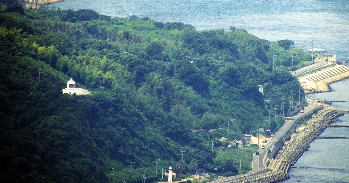 從明石海峽大橋主塔往下看淡路島北端的野島江崎一帶:長滿樹林的山坡上立著白色的江崎燈塔,山腳下一條沿海道路貼著消波塊海堤,右邊是藍色海面