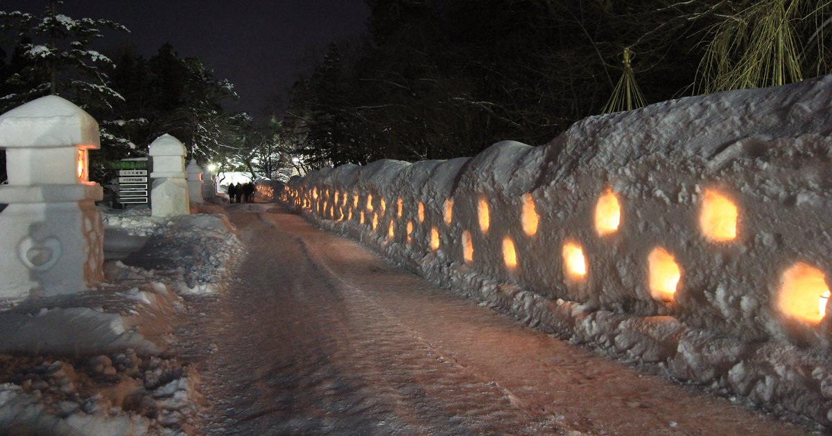 弘前公園雪燈籠祭夜景:右側一整道雪牆挖出一排排方形小洞,裡面點著蠟燭發出橘光,左側立著幾座雪做的燈籠,中間是壓實的雪地步道,遠處有幾個散步的人影