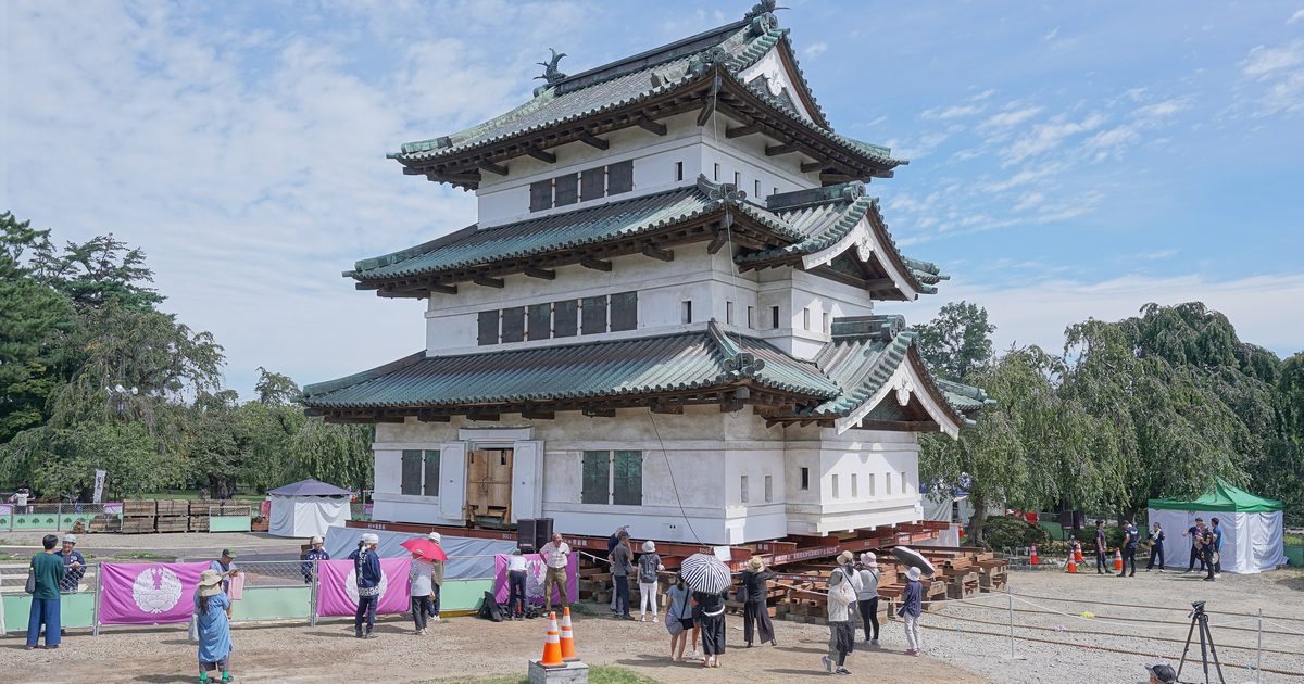 Hirosaki Castle keep jacked up whole and resting on red steel beams over stacked timber cribbing on gravel, ringed by purple curtains bearing the Tsugaru crest and a crowd of onlookers