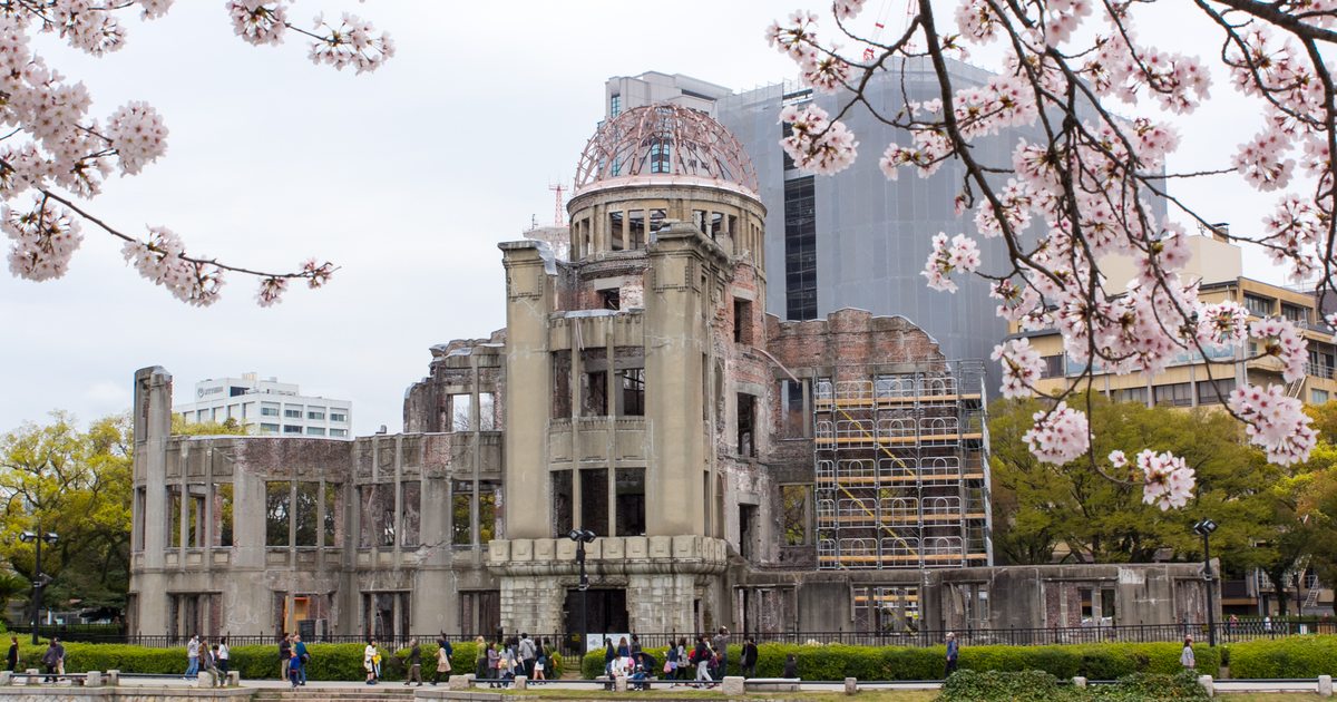2016 年 4 月從平和記念公園一側隔著元安川看原爆圓頂,前景左右兩側垂著盛開的櫻花枝條,圓頂右側搭著保存工程的鷹架,對岸步道上有行人
