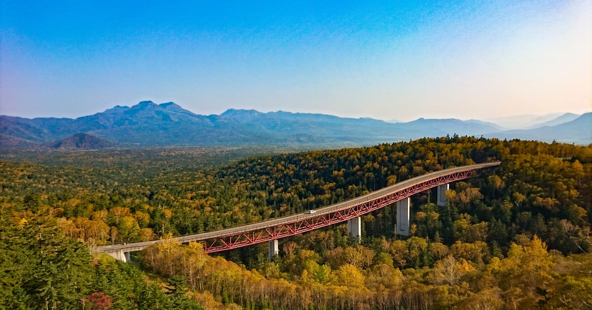 Hokkaido Mikuni Pass autumn road from above: S-curve mountain highway flanked by orange-gold foliage and distant Daisetsuzan range