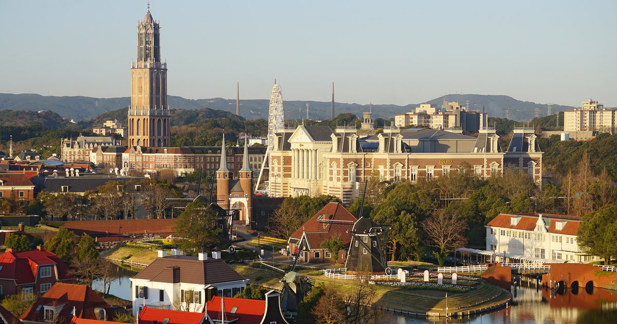 Elevated view over Huis Ten Bosch: the Domtoren replica at left, a classical brick facade at centre, black windmills and red-tiled Dutch houses along the canal