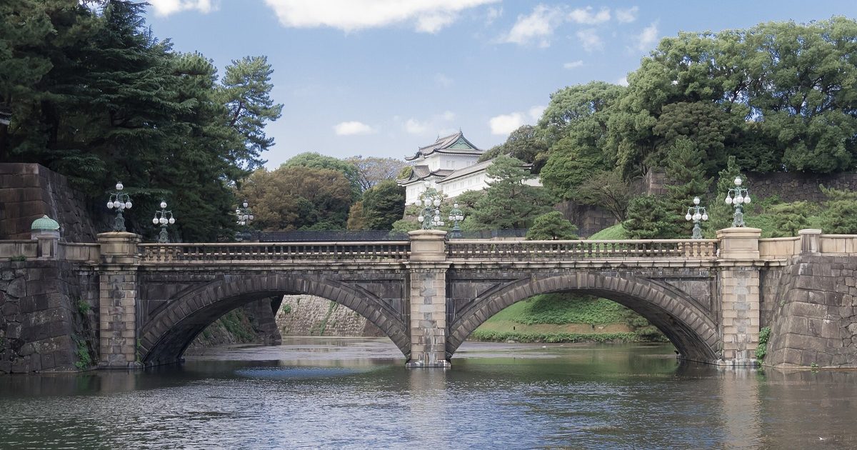 從皇居外苑隔著護城河看皇居正門石橋：雙拱石橋橫過畫面，橋上立著青銅燈柱，後方樹林間露出白牆黑瓦的伏見櫓