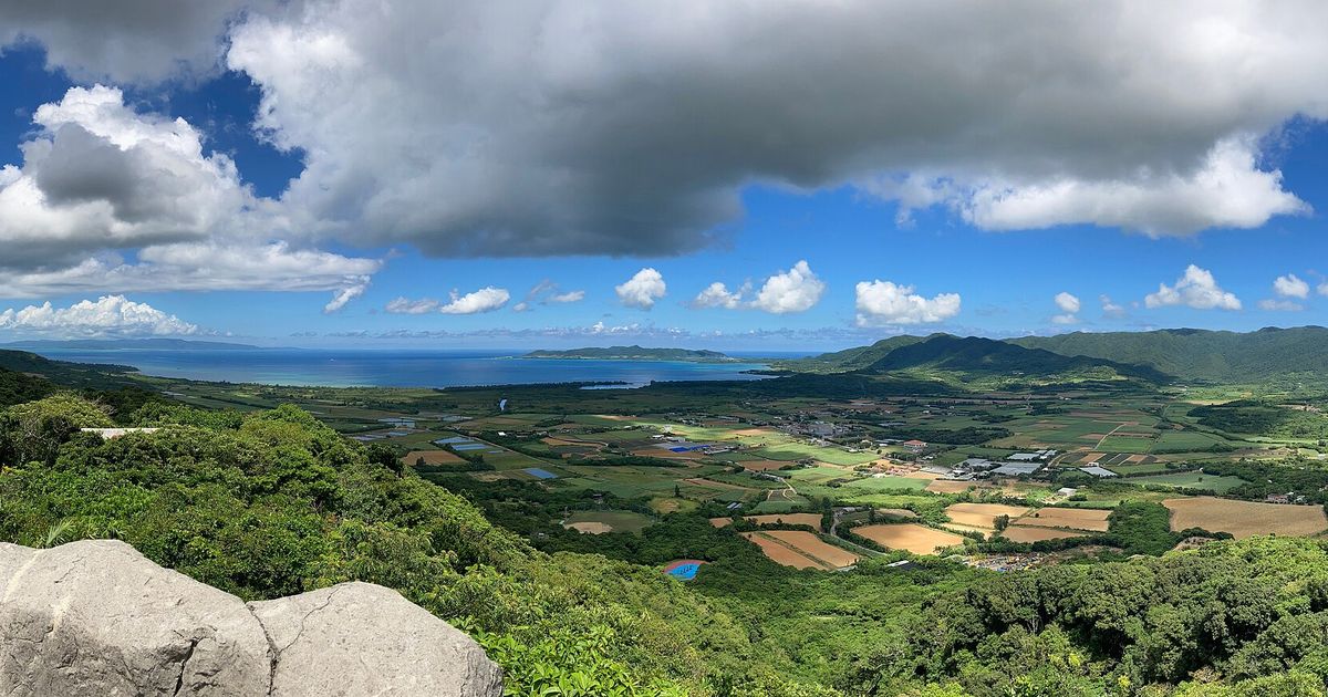 從バンナ公園展望台往西俯瞰石垣島:前景是綠色山林,中間一大片田地與零星房舍的名蔵平原,遠方是藍色的名蔵湾與崎枝半島,右側是於茂登岳一帶的山,天上有大片積雲