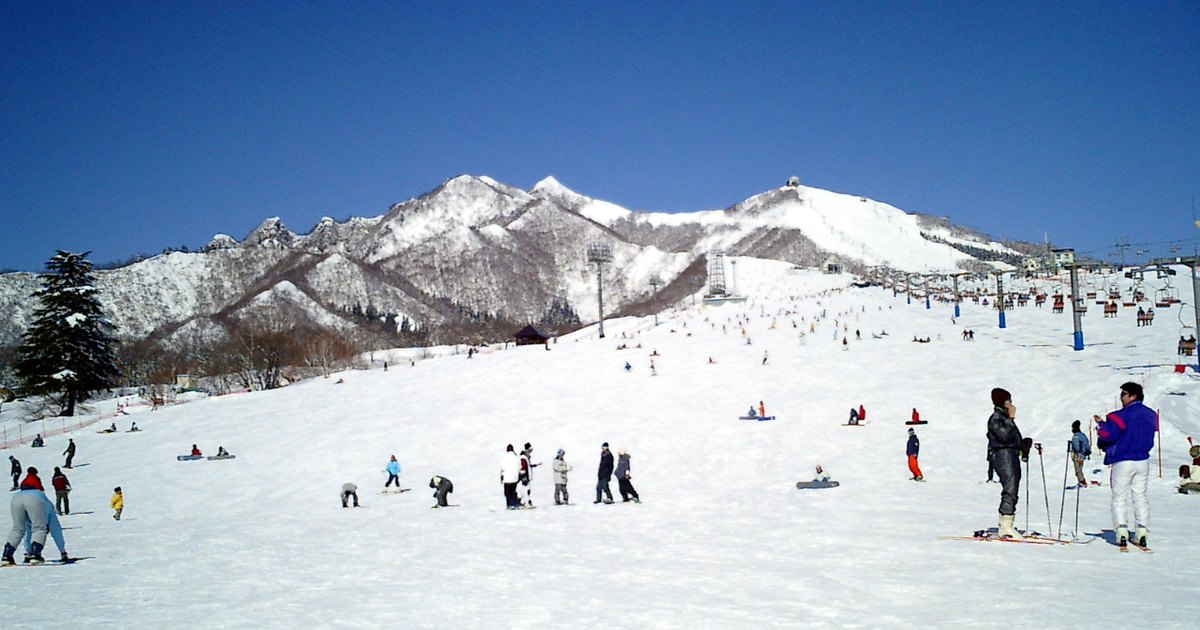晴天的岩原滑雪場山腳:寬闊平緩的壓雪雪道往上延伸,右側一整排吊椅纜車,坡上立著高高的照明燈柱,後方是覆雪的飯士山一帶山稜;雪道上散著滑雪和單板的人。2004 年 2 月拍攝