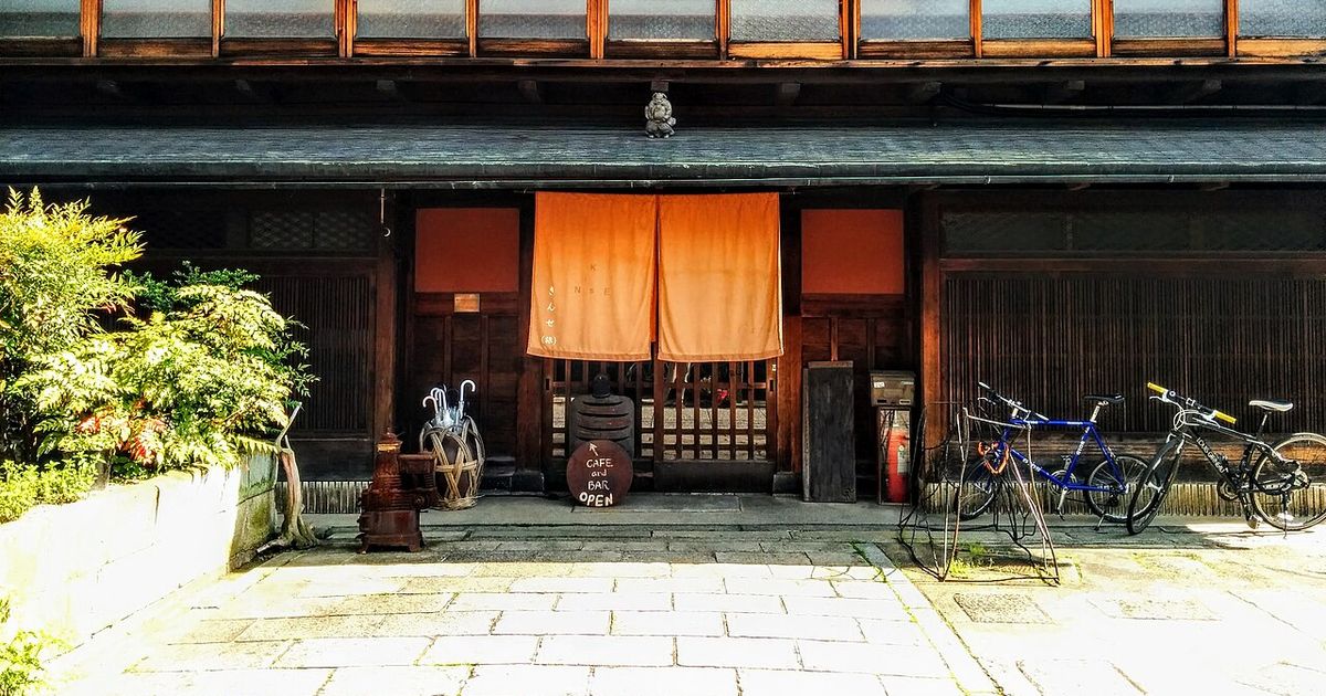Wooden facade of a Kyoto machiya inn: a row of latticed glass windows upstairs, an orange noren curtain hanging over the ground-floor entrance, a round chalkboard reading CAFE and BAR beside it, two bicycles parked to the right and stone paving in front