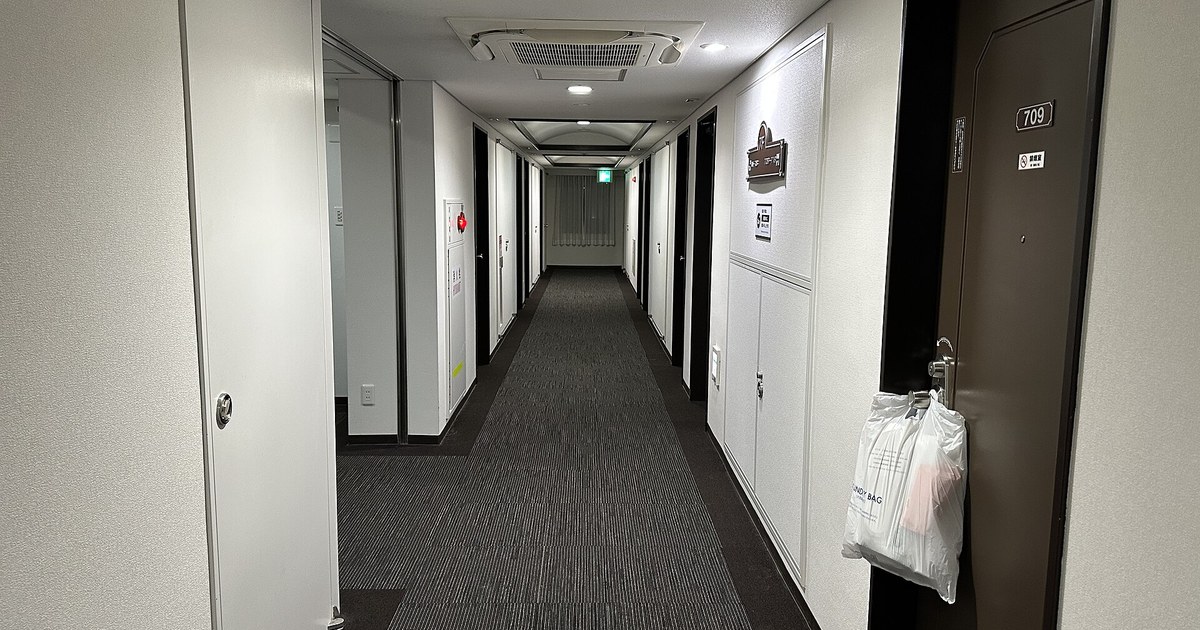 A Japanese business hotel guest floor corridor, white walls and dark brown room doors, a laundry bag hanging on the handle of room 709, a curtained window at the far end