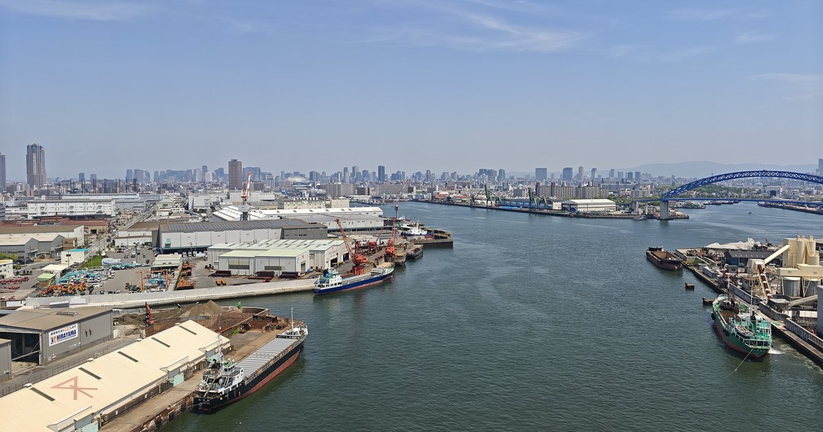 Osaka's port waterway seen from a bridge: white warehouses, cranes and moored cargo ships line both banks, a row of city towers spans the far horizon, a blue steel arch bridge stands to the right, and the sky is clear enough to show the mountain ridge behind the city