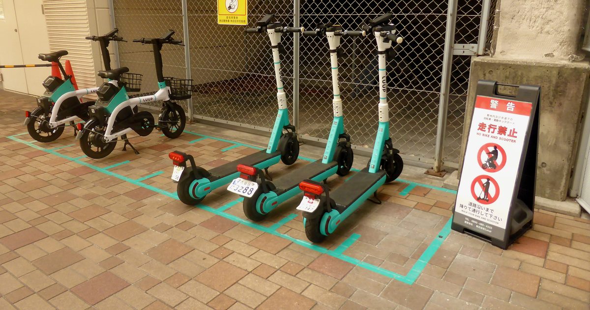 Parking bays marked out in turquoise tape on a brick pavement, with three white-and-turquoise electric kick scooters lined up inside them, each carrying a small number plate and a red rear lamp; two seated machines of the same livery stand behind against a chain-link fence, and a red-and-white A-frame board to the right reads NO BIKE AND SCOOTER