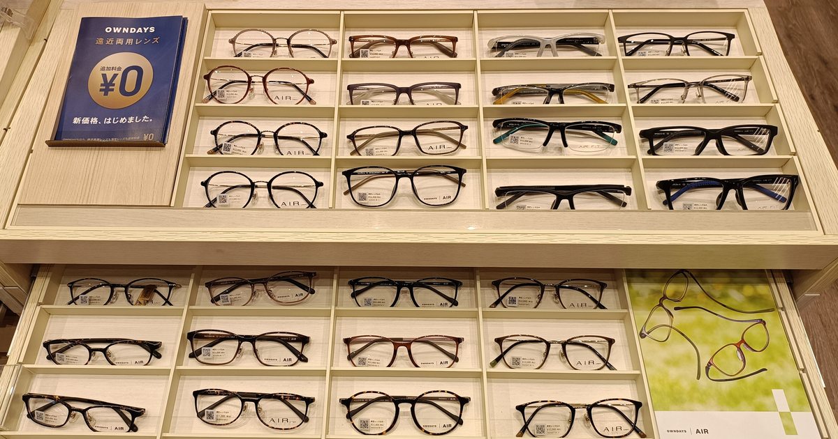 Frame trays at an OWNDAYS store in Japan, each frame in its own compartment with a price tag, beside a card advertising progressive lenses