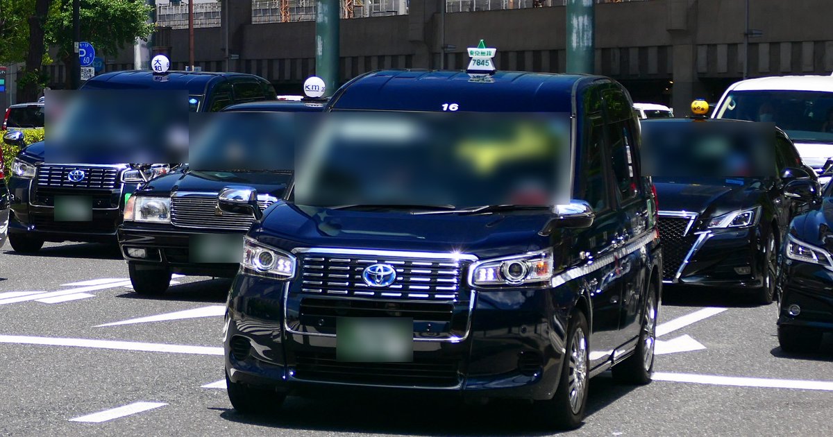 東京車站丸之內側的路口，幾台深藍色的 JPN TAXI 計程車與一台黑色轎車型計程車排在斑馬線前，背景是高架鐵道
