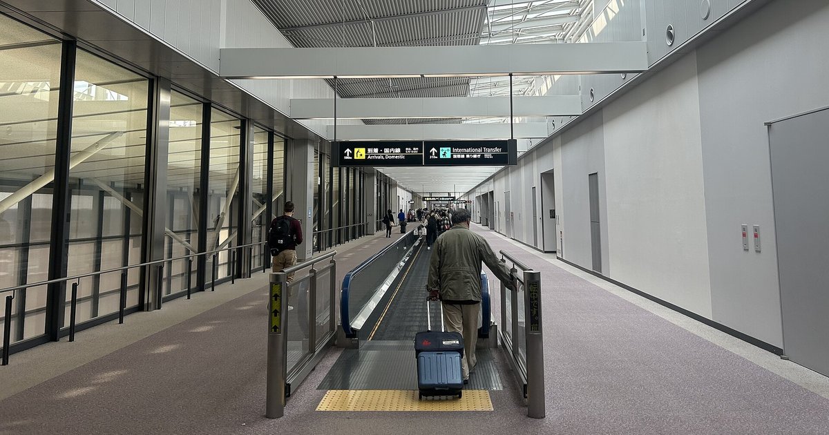 成田機場第 1 航廈的國際線到達通道：旅客拖著行李箱站上電動步道，上方指標寫著「到着・国内線 Arrivals」與「International Transfer」