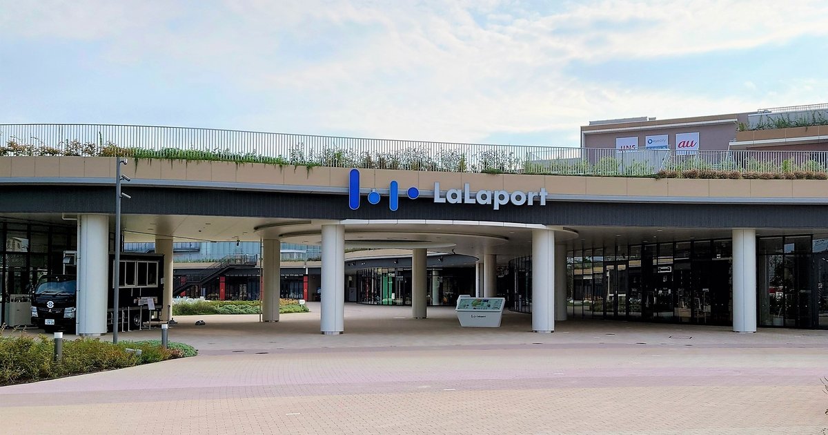LaLaport 福岡的フラワーパーク側入口:米色屋簷上掛著藍色的 LaLaport 標誌,下方一排白色圓柱撐起弧形的入口通道,屋頂是種滿植物的空中步道,前方是空曠的地磚廣場