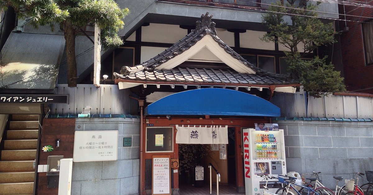 The street frontage of Tsubameyu, a sento in Ueno, Tokyo: a tiled curved gable over the doorway, a white noren curtain hanging in the entrance, a blue awning and a plaque listing the opening hours
