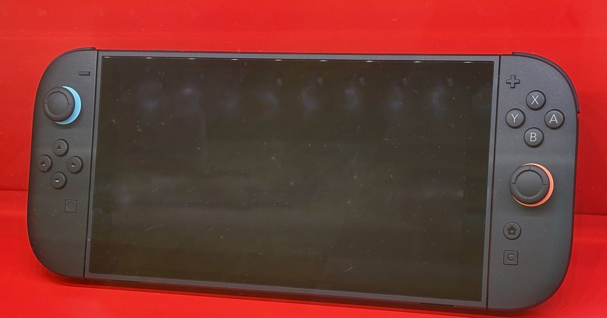 紅色展示櫃裡的 Nintendo Switch 2 主機：黑色螢幕兩側接著深灰色的 Joy-Con 2，左搖桿底下一圈藍色、右搖桿底下一圈橘色，右下角有方形的 C 鍵