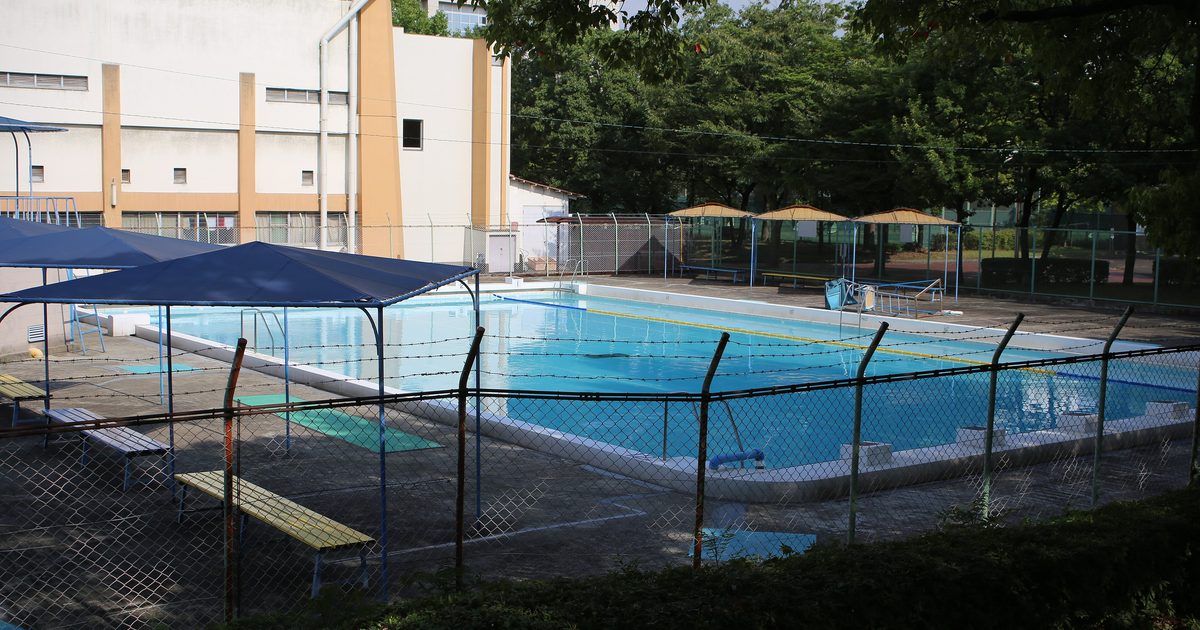 A Japanese municipal outdoor pool in summer: a rectangular pool filled with still, empty water, blue shade canopies and benches along the deck, four thatched parasols at the far end, chain-link fencing around the whole area, and a cream sports hall and trees behind
