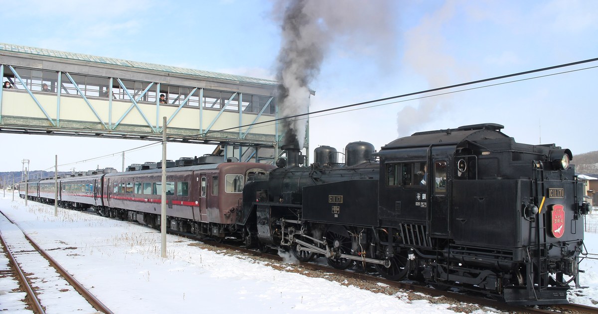 A Japanese winter sightseeing train: a steam locomotive carrying the SL Fuyu no Shitsugen headboard, maroon coaches behind it, standing at a snow-covered station under a blue sky