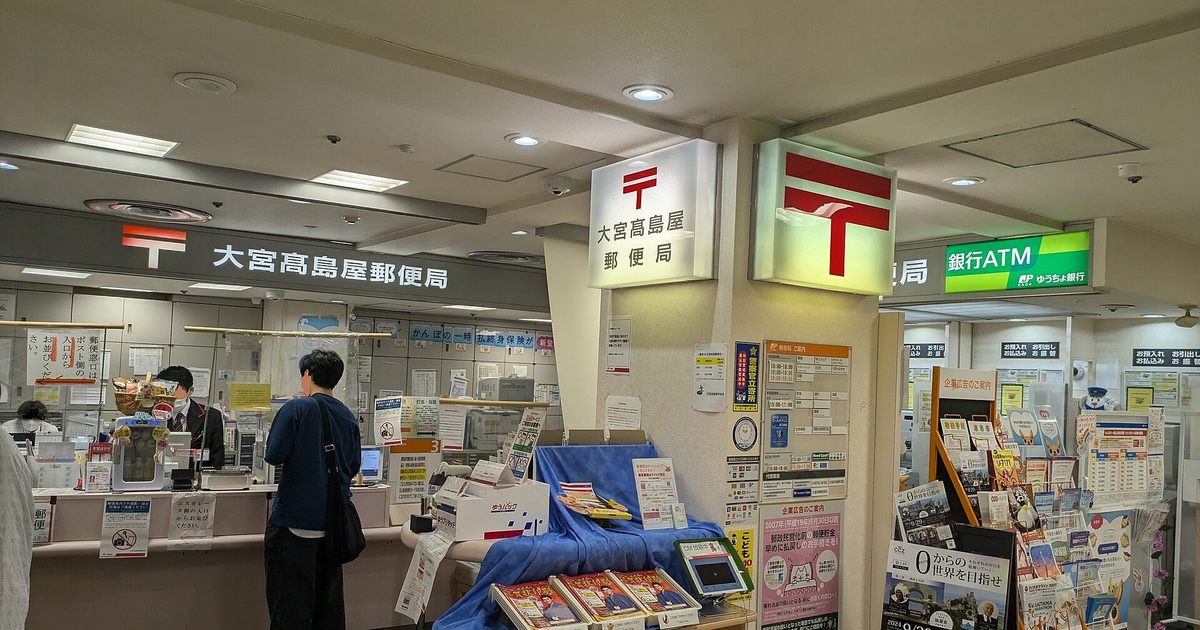 The service floor of Omiya Takashimaya Post Office in Japan: two illuminated red postal-mark signs above a long counter where a uniformed clerk serves a customer with a shoulder bag, a Yu-Pack carton and printed leaflets on the table in the middle, and the Japan Post Bank ATM area with racks of brochures on the right