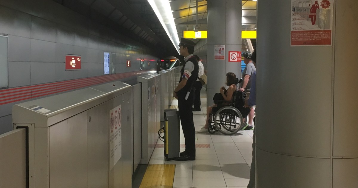 An underground station platform: a uniformed attendant waits by the platform gate with a portable boarding ramp at hand, while a passenger in a manual wheelchair waits beside a pillar with companions; yellow tactile paving runs along the edge and a train’s headlights approach down the tunnel